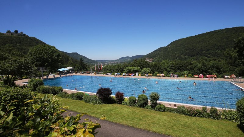 Ein Freibad inmitten einer gr&uuml;nen, bergigen Landschaft. Menschen schwimmen im gro&szlig;en Becken, umgeben von B&auml;umen und H&uuml;geln unter klarem Himmel.