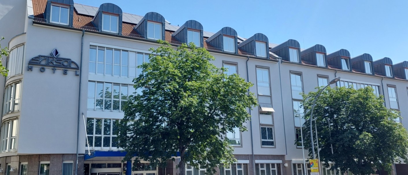 Moderne Hotelfassade mit großen Fenstern und Dachgauben, ein Baum und ein Auto davor. Klare, sonnige Wetterbedingungen., © Erikson Hotel Moderne Hotelfassade mit großen Fenstern und Dachgauben, ein Baum und ein Auto davor. Klare, sonnige Wetterbedingungen., © Erikson Hotel