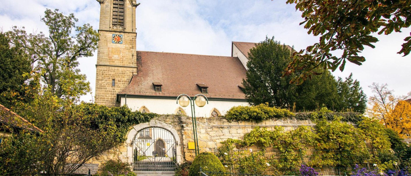 Eine historische Kirche in Leinfelden-Echterdingen, umgeben von üppigem Grün und herbstlichen Bäumen. Ein Tor führt zum Eingang der Kirche., © Stuttgart-Marketing GmbH, Sarah Schmid