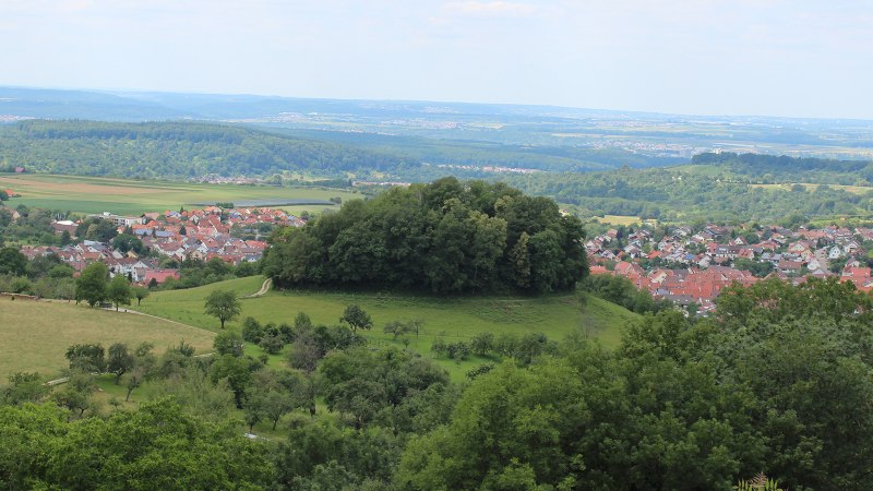 Blick auf Beuren mit einem bewaldeten H&uuml;gel im Vordergrund und einem Dorf mit roten D&auml;chern im Hintergrund, umgeben von gr&uuml;ner Landschaft., &copy; Kurverwaltung Beuren