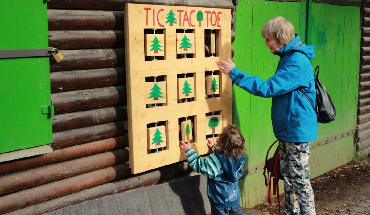 Ein Kind und eine ältere Person spielen Tic Tac Toe an einer Holzwand mit grünen Symbolen. Sie stehen vor einer Hütte mit grünen Türen., © Amt für Forsten Landkreis Böblingen Ein Kind und eine ältere Person spielen Tic Tac Toe an einer Holzwand mit grünen Symbolen. Sie stehen vor einer Hütte mit grünen Türen., © Amt für Forsten Landkreis Böblingen