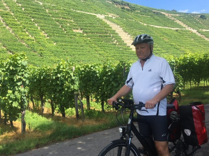 Ein Mann mit Helm steht mit einem E-Bike vor grünen Weinbergen. Er trägt ein weißes Trikot und hat eine rote Fahrradtasche., © Weingenuss Stuttgart Ein Mann mit Helm steht mit einem E-Bike vor grünen Weinbergen. Er trägt ein weißes Trikot und hat eine rote Fahrradtasche., © Weingenuss Stuttgart