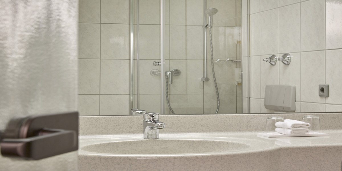 Ein modernes Badezimmer mit einem Waschbecken, einem Spiegel und einer Dusche mit Glastür. Handtücher und Gläser sind auf dem Waschbecken platziert., © © H-Hotels.com