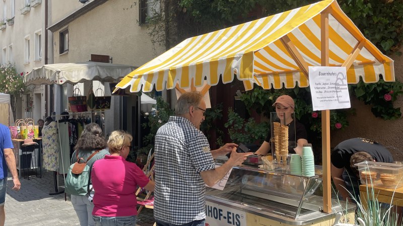 Ein Eiswagen mit gelb-wei&szlig;em Sonnendach steht in einer schmalen Gasse. Menschen kaufen Eis. Die Umgebung ist von Pflanzen und alten Geb&auml;uden gepr&auml;gt., &copy; Stadt Schorndorf