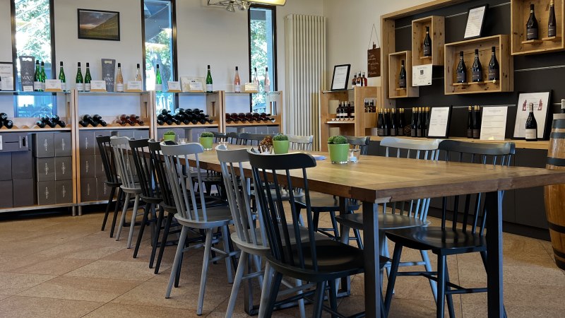 Vinothek mit Holztisch und Stühlen, umgeben von Weinregalen und Dekoration. Helle, einladende Atmosphäre mit Weinflaschen und Pflanzen., © Weinfactum Bad Cannstatt