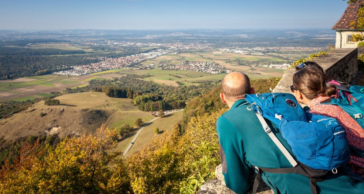 Zwei Personen mit Rucksäcken blicken von der Burg Teck auf eine weite Landschaft mit Feldern und Dörfern unter blauem Himmel., © hochgehberge Zwei Personen mit Rucksäcken blicken von der Burg Teck auf eine weite Landschaft mit Feldern und Dörfern unter blauem Himmel., © hochgehberge