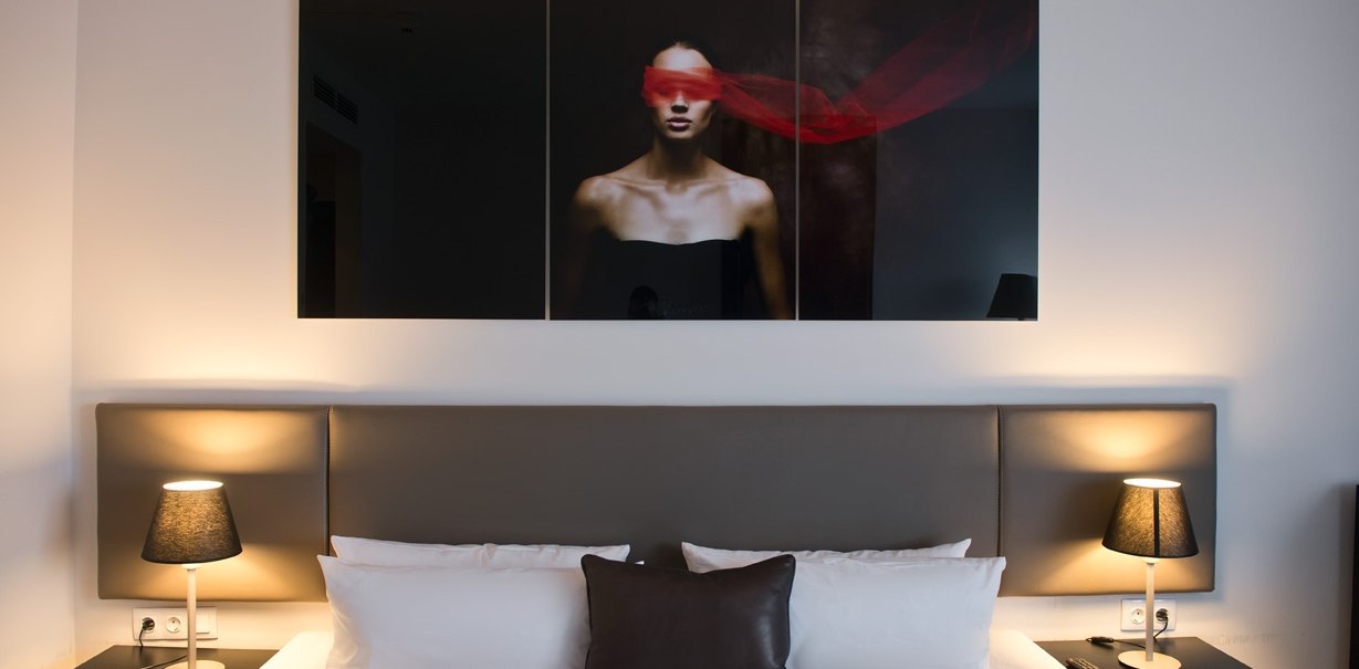 Modernes Hotelzimmer mit Doppelbett, zwei Nachttischlampen und einem Kunstwerk über dem Bett, das eine Person mit rotem Tuch zeigt., © Wyndham Stuttgart Airport Messe