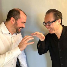 Zwei Männer in Hemden lächeln sich an und heben spielerisch die Hände, als ob sie Klavier spielen., © links im Bild Zwei Männer in Hemden lächeln sich an und heben spielerisch die Hände, als ob sie Klavier spielen., © links im Bild