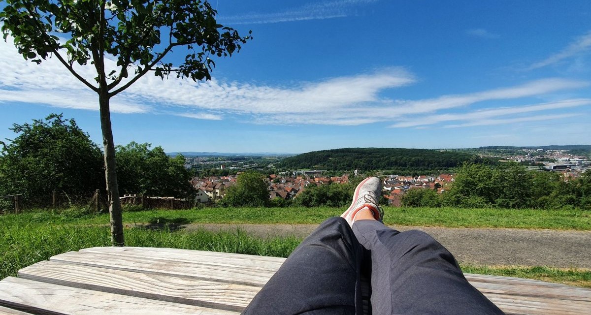 Person entspannt auf einer Holzbank mit Blick auf eine Stadtlandschaft und grüne Hügel unter blauem Himmel. Person entspannt auf einer Holzbank mit Blick auf eine Stadtlandschaft und grüne Hügel unter blauem Himmel.