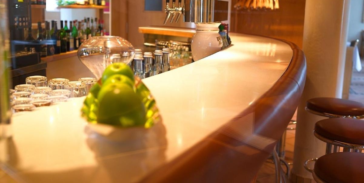 Elegante Bar mit geschwungener Theke, Barhockern und Flaschen im Hintergrund. Im Vordergrund liegen gr&uuml;ne &Auml;pfel auf einem Teller., &copy; Hotel am Park