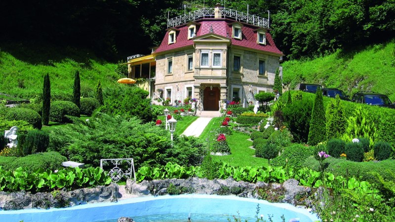 Ein kleines Schloss mit rotem Dach, umgeben von einem gepflegten Garten mit Springbrunnen und üppiger Vegetation. Ein kleines Schloss mit rotem Dach, umgeben von einem gepflegten Garten mit Springbrunnen und üppiger Vegetation.