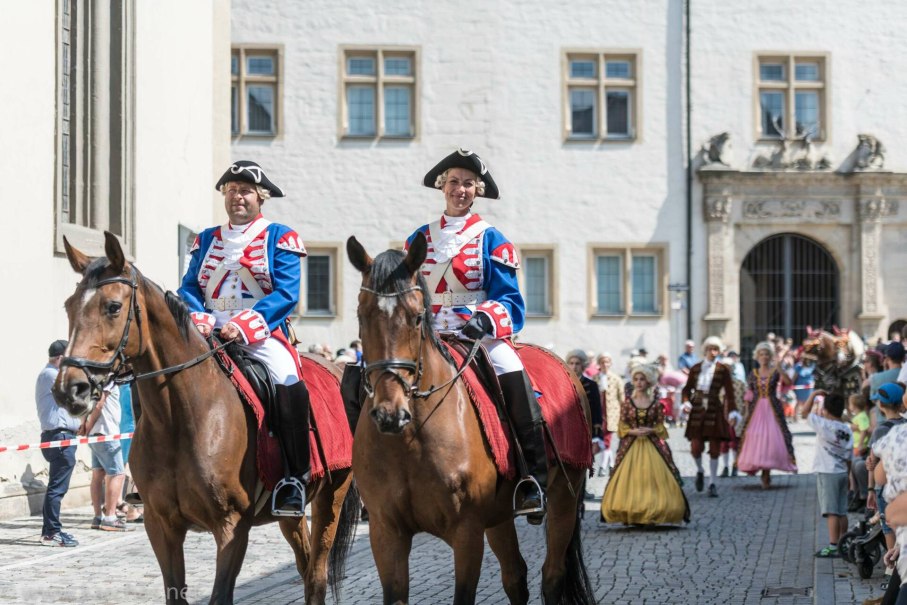 Zwei Reiter in blauen historischen Uniformen auf Pferden bei einem Umzug. Im Hintergrund sind weitere kostümierte Personen und Zuschauer zu sehen.