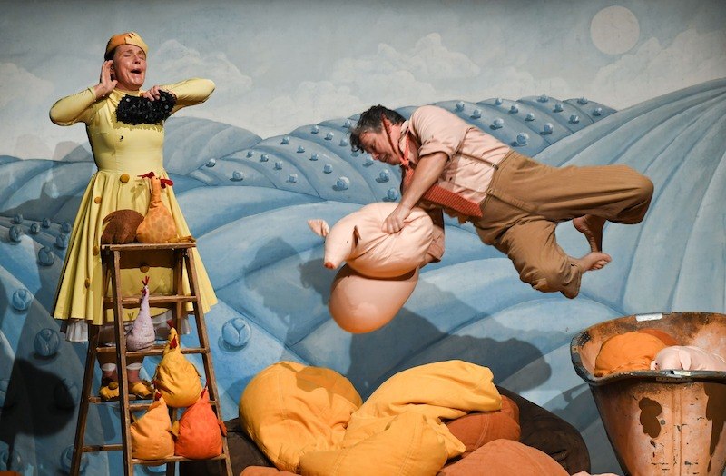 Bühnenbild mit zwei Schauspielern: Einer springt mit einem Stoffschwein, der andere steht auf einer Leiter. Bunte Hügel und Stofftiere im Hintergrund., © Stadt Kornwestheim Kulturmanagement
