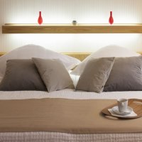 Modernes Schlafzimmer mit Doppelbett, dekoriert mit Kissen und Tablett mit Tasse. Zwei rote Vasen auf einem Regal im Hintergrund., © Hotel Drei Morgen Modernes Schlafzimmer mit Doppelbett, dekoriert mit Kissen und Tablett mit Tasse. Zwei rote Vasen auf einem Regal im Hintergrund., © Hotel Drei Morgen