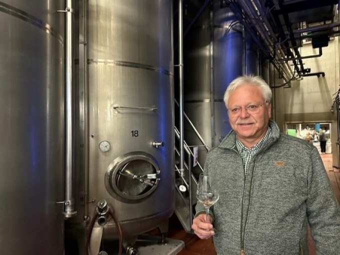 Ein Mann in einer Weinkellerei hält ein Weinglas vor großen Edelstahltanks. Die Umgebung wirkt industriell und sauber., © Weingenuss Stuttgart Ein Mann in einer Weinkellerei hält ein Weinglas vor großen Edelstahltanks. Die Umgebung wirkt industriell und sauber., © Weingenuss Stuttgart