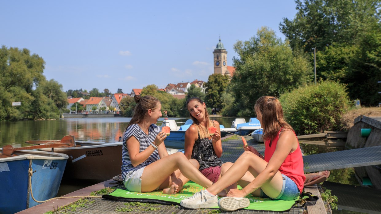 Drei Frauen sitzen auf einem Steg am Fluss und essen Wassermelone. Im Hintergrund sind Boote und ein Kirchturm zu sehen., © H. Bergmüller Drei Frauen sitzen auf einem Steg am Fluss und essen Wassermelone. Im Hintergrund sind Boote und ein Kirchturm zu sehen., © H. Bergmüller