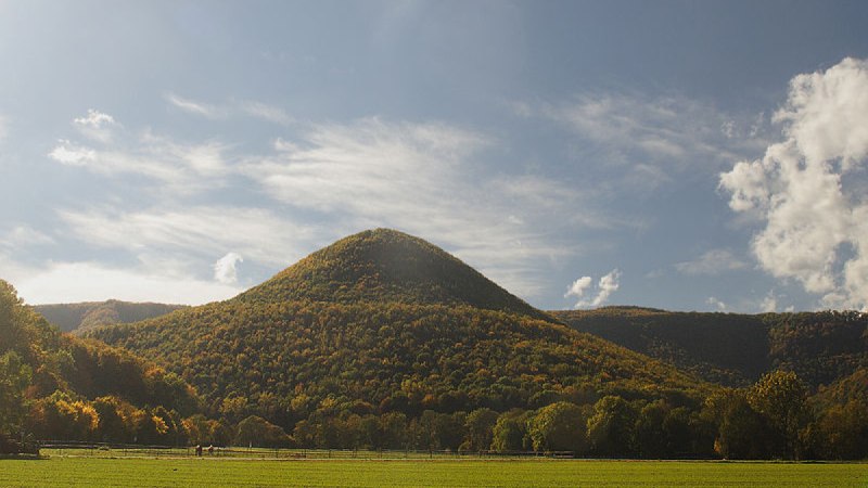 Ein runder, bewaldeter Berg erhebt sich unter einem blauen Himmel mit Wolken. Im Vordergrund sind gr&uuml;ne Felder und B&auml;ume zu sehen., &copy; Bad Urach Tourismus
