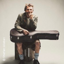 Ein Mann sitzt lächelnd auf einem Holzkasten, lehnt sich auf einen Gitarrenkoffer. Er trägt eine Jacke und Socken mit Schuhen., © links im Bild