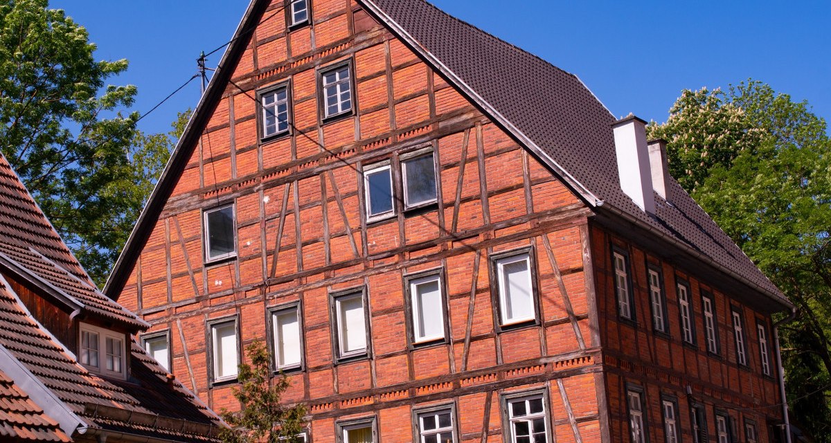 Ein großes Fachwerkhaus mit rotem Ziegelmauerwerk und dunklem Dach steht vor einem klaren blauen Himmel. Bäume umgeben das Gebäude., © Agentur Arcos Ein großes Fachwerkhaus mit rotem Ziegelmauerwerk und dunklem Dach steht vor einem klaren blauen Himmel. Bäume umgeben das Gebäude., © Agentur Arcos