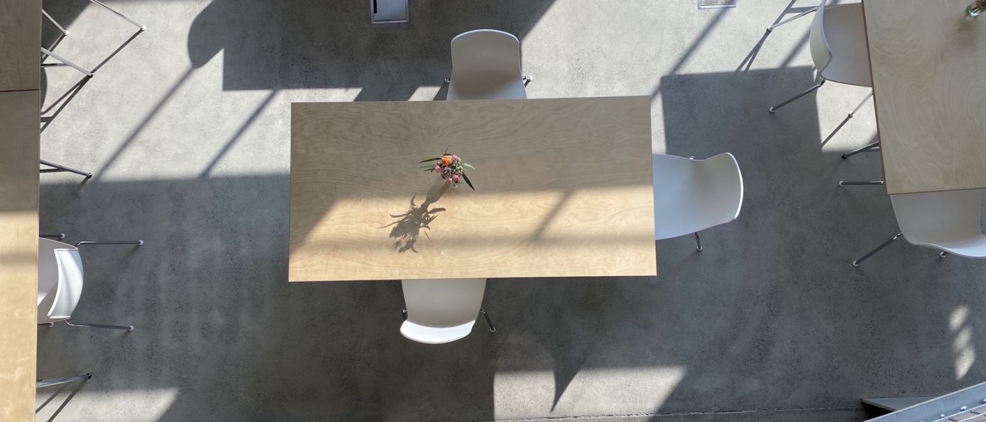 Luftaufnahme eines Seminarraums mit Holztischen und weißen Stühlen. Sonnenlicht wirft Schatten auf den Betonboden. Eine Blumenvase steht auf einem Tisch., © Nina Kiesel Luftaufnahme eines Seminarraums mit Holztischen und weißen Stühlen. Sonnenlicht wirft Schatten auf den Betonboden. Eine Blumenvase steht auf einem Tisch., © Nina Kiesel