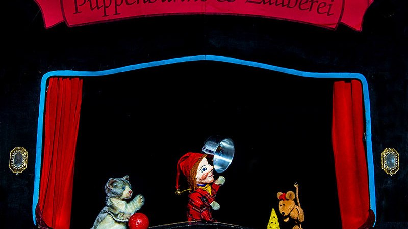 Eine Puppenbühne zeigt eine Katze, eine Puppe mit einem Topf und eine Maus mit Käse. Der Vorhang ist rot, der Hintergrund schwarz., © Prinzessin Gisela Theater Eine Puppenbühne zeigt eine Katze, eine Puppe mit einem Topf und eine Maus mit Käse. Der Vorhang ist rot, der Hintergrund schwarz., © Prinzessin Gisela Theater