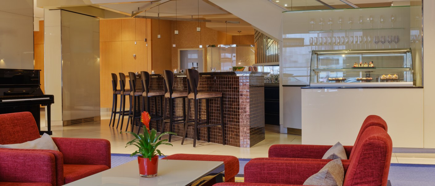 Moderne Hotellobby mit roten Sesseln, einem Tisch mit Pflanze, einer Kuchenvitrine und einer Bar mit hohen Stühlen., © Steigenberger Graf Zeppelin