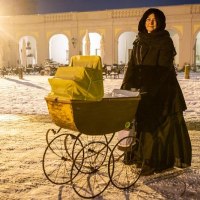 Eine Person in schwarzem Mantel steht mit einem antiken Kinderwagen im Schnee vor einem beleuchteten Gebäude mit Bögen., © Tourist Information und Ticket Service Ludwigsburg