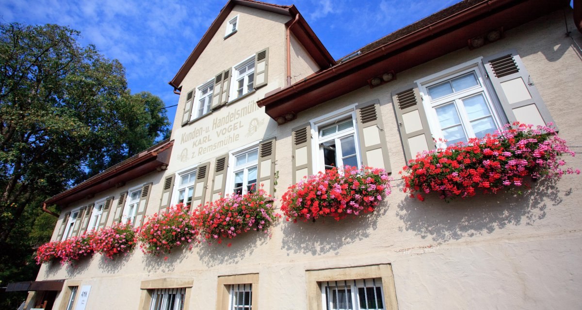 Historisches Geb&auml;ude mit wei&szlig;en Fensterl&auml;den und bl&uuml;henden Blumenk&auml;sten vor einem blauen Himmel. Schriftzug "Kunden- u. Handelsm&uuml;hle Karl Vogel z. Remsm&uuml;hle" sichtbar., &copy; Remstal Tourismus e.V.