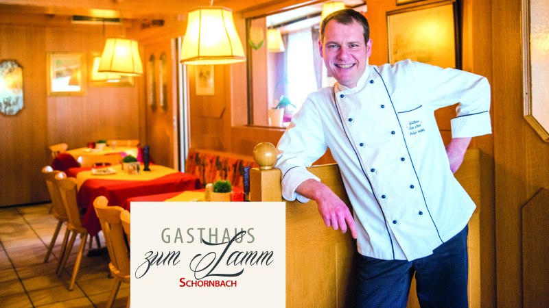 Ein Koch in weißer Uniform steht lächelnd in einem gemütlichen Gasthaus mit Holzmöbeln und roten Tischdecken., © Gasthaus Lamm Schornbach, Fotografie Nicole Schielberg