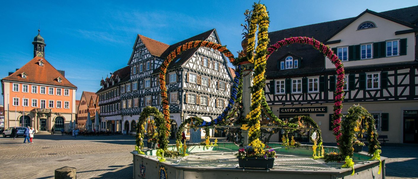 Der Marktplatz in Schorndorf zeigt einen geschmückten Brunnen vor historischen Fachwerkhäusern und dem Rathaus. Menschen spazieren über den Platz., © Stuttgart-Marketing GmbH, Sarah Schmid Der Marktplatz in Schorndorf zeigt einen geschmückten Brunnen vor historischen Fachwerkhäusern und dem Rathaus. Menschen spazieren über den Platz., © Stuttgart-Marketing GmbH, Sarah Schmid
