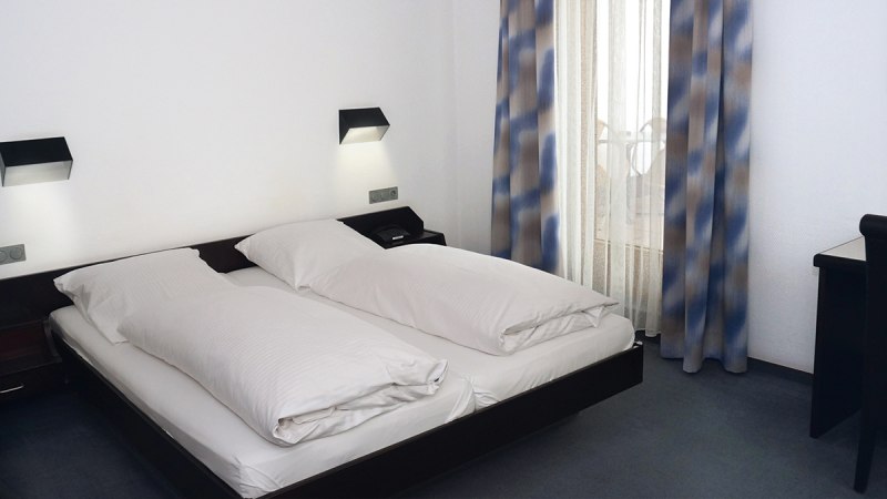 Ein schlichtes Hotelzimmer mit Doppelbett, weißen Bettdecken und blau-braunen Vorhängen. An der Wand sind zwei Leselampen angebracht., © TOMAS Ein schlichtes Hotelzimmer mit Doppelbett, weißen Bettdecken und blau-braunen Vorhängen. An der Wand sind zwei Leselampen angebracht., © TOMAS