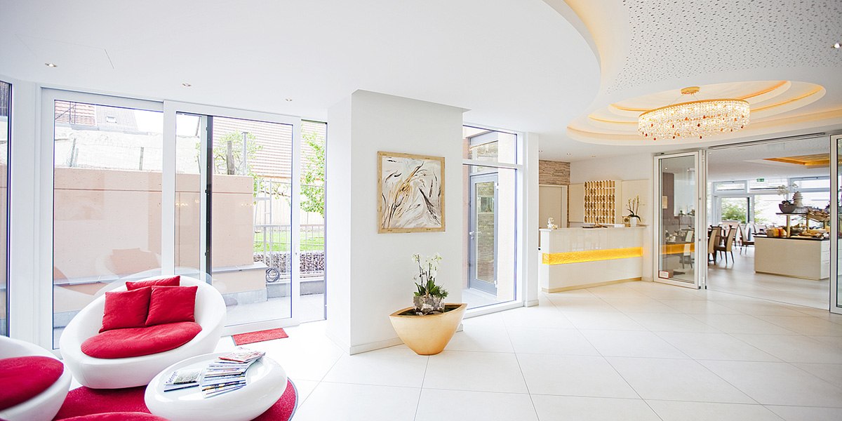 Helle Lobby mit modernen weißen Möbeln, roten Kissen, großem Kronleuchter und Glaswänden. Empfangsbereich mit Pflanzen und Kunstwerk an der Wand., © Hotel Krone Helle Lobby mit modernen weißen Möbeln, roten Kissen, großem Kronleuchter und Glaswänden. Empfangsbereich mit Pflanzen und Kunstwerk an der Wand., © Hotel Krone