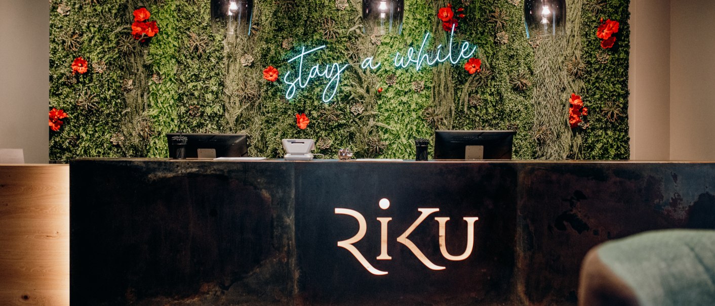Rezeption mit grünem Pflanzenhintergrund, roten Blumen und Leuchtschrift 'stay a while'. Der Tresen trägt das Logo 'RiKU'., © 13 6 Design GmbH (Judenhof 10, 89073 Ulm) Rezeption mit grünem Pflanzenhintergrund, roten Blumen und Leuchtschrift 'stay a while'. Der Tresen trägt das Logo 'RiKU'., © 13 6 Design GmbH (Judenhof 10, 89073 Ulm)