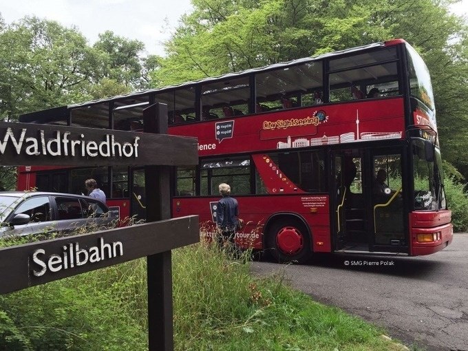 Roter Doppeldeckerbus auf einer grünen Straße, Wegweiser zum Waldfriedhof und zur Seilbahn im Vordergrund., © SMG, Pierre Polak Roter Doppeldeckerbus auf einer grünen Straße, Wegweiser zum Waldfriedhof und zur Seilbahn im Vordergrund., © SMG, Pierre Polak