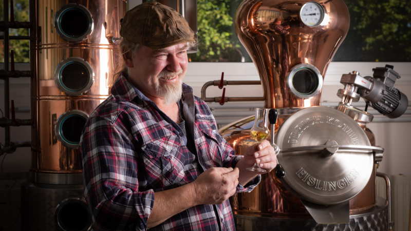 Mann in kariertem Hemd h&auml;lt ein Glas mit goldenem Getr&auml;nk vor einer Kupferdestille. Er l&auml;chelt und tr&auml;gt eine M&uuml;tze. Im Hintergrund sind Fenster und B&auml;ume sichtbar., &copy; Brau- & Brennwerkstatt