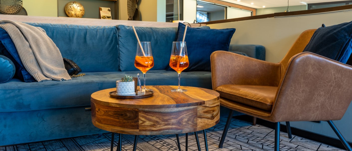 Gemütliche Lobby mit blauer Couch, braunem Sessel und Holztisch. Zwei Aperol Spritz Gläser stehen auf dem Tisch, im Hintergrund Regale mit Dekoration., © MASEVEN Stuttgart SI-Centrum Gemütliche Lobby mit blauer Couch, braunem Sessel und Holztisch. Zwei Aperol Spritz Gläser stehen auf dem Tisch, im Hintergrund Regale mit Dekoration., © MASEVEN Stuttgart SI-Centrum