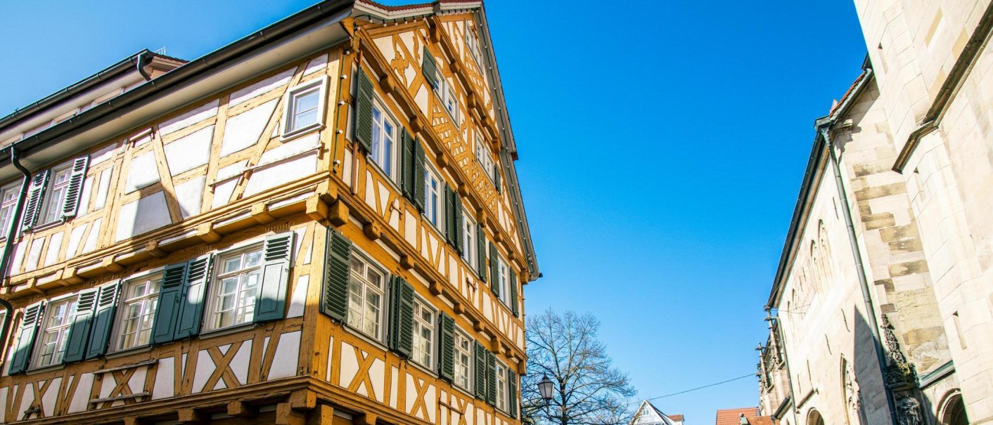 Ein Fachwerkhaus in Schorndorf mit grünen Fensterläden, daneben ein historisches Gebäude. Der Himmel ist klar und blau., © Stuttgart-Marketing GmbH, Sarah Schmid