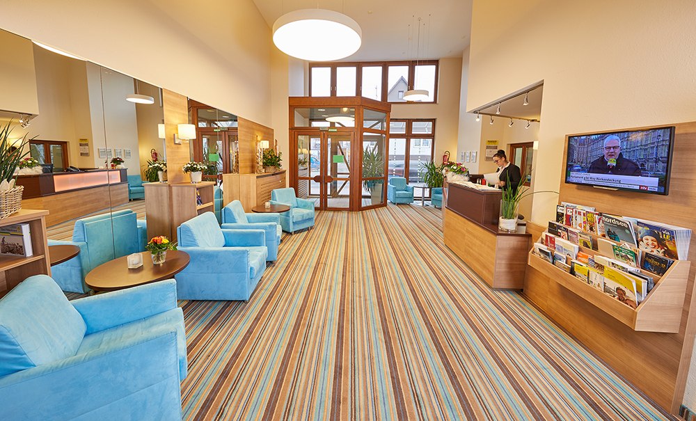 Helle Lobby mit blauen Sesseln, Holzmöbeln und gestreiftem Teppich. Rezeption mit Mitarbeiter, TV an der Wand, Zeitschriftenständer., © TOMAS