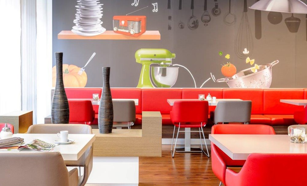 Moderner Frühstücksbereich mit roten und grauen Stühlen, dekorativen Vasen und einer Wand mit Küchenmotiven., © Ibis Stuttgart Airport Messe Moderner Frühstücksbereich mit roten und grauen Stühlen, dekorativen Vasen und einer Wand mit Küchenmotiven., © Ibis Stuttgart Airport Messe