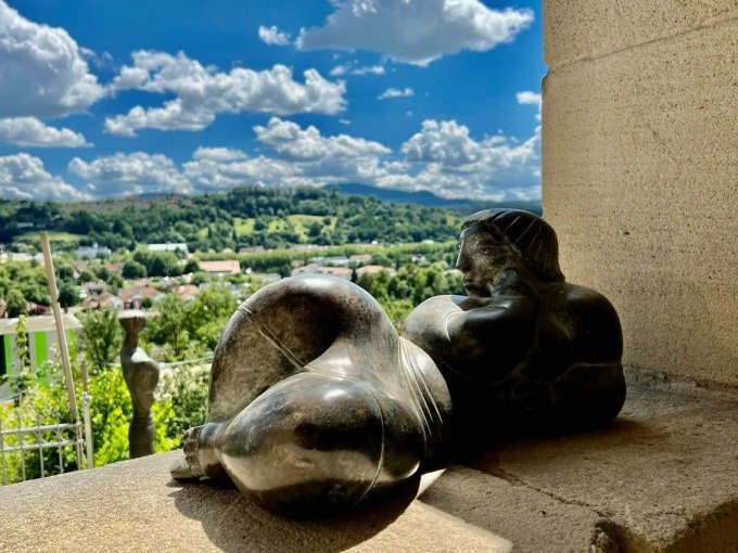 Eine Skulptur einer liegenden Frau auf einer Terrasse, die auf eine grüne Landschaft und einen blauen Himmel mit Wolken blickt., © Touristik und Marketing GmbH Eine Skulptur einer liegenden Frau auf einer Terrasse, die auf eine grüne Landschaft und einen blauen Himmel mit Wolken blickt., © Touristik und Marketing GmbH
