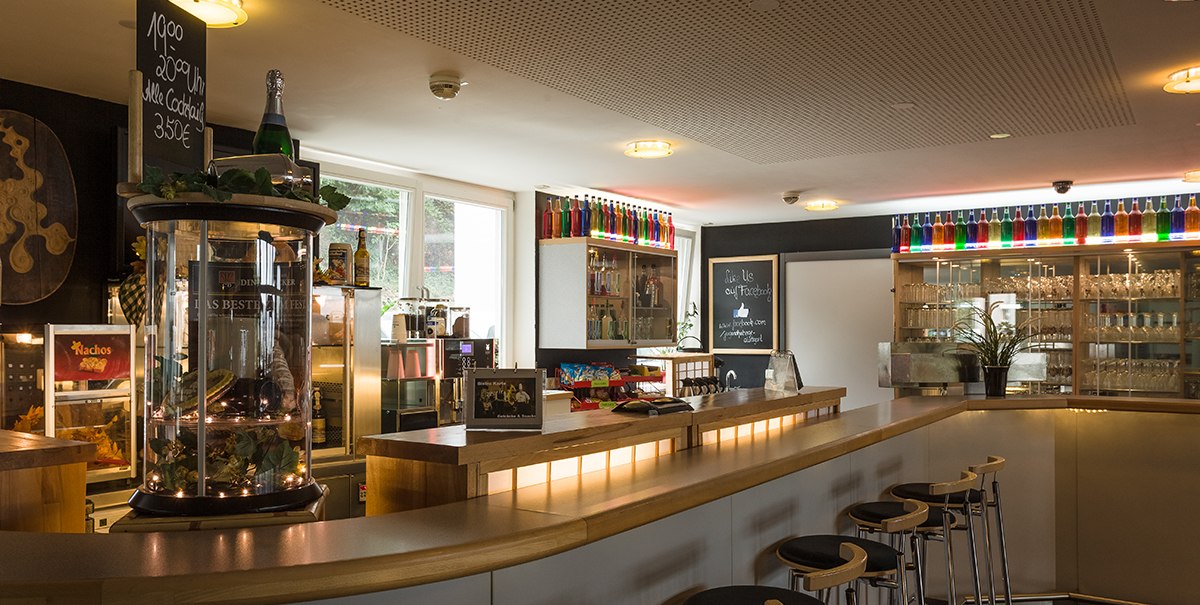 Ein gemütliches Bistro mit einer langen Theke, Barhockern und dekorativen Flaschen auf Regalen. Ein Schild bewirbt Cocktails., © TOMAS Ein gemütliches Bistro mit einer langen Theke, Barhockern und dekorativen Flaschen auf Regalen. Ein Schild bewirbt Cocktails., © TOMAS