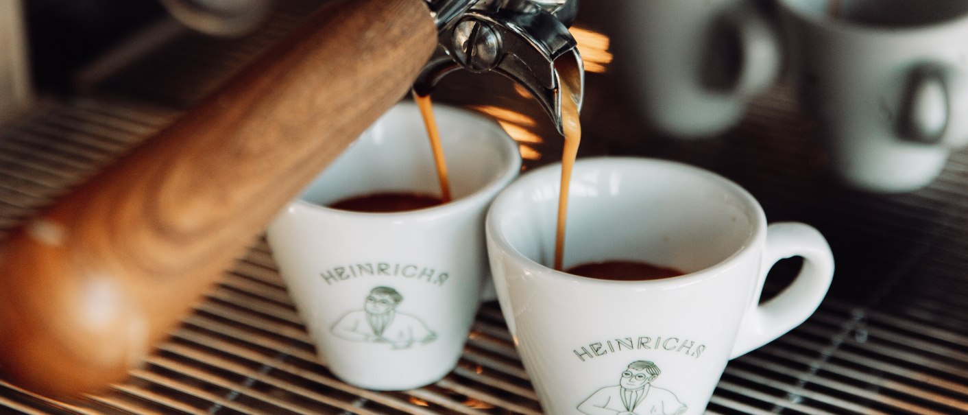 Eine Espressomaschine gießt Kaffee in zwei Tassen mit dem Logo von Heinrichs Cafe. Die Tassen stehen auf einem Metallgitter., © L. Prestle/ Documenting Love Eine Espressomaschine gießt Kaffee in zwei Tassen mit dem Logo von Heinrichs Cafe. Die Tassen stehen auf einem Metallgitter., © L. Prestle/ Documenting Love