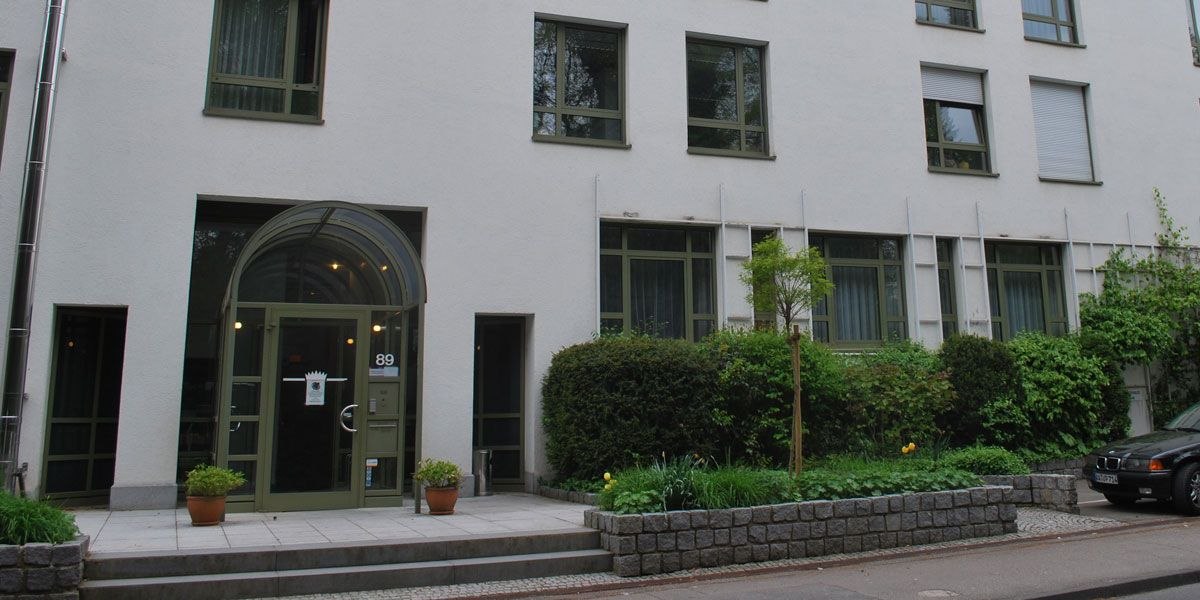 Moderner Gebäudeeingang mit Glasdach und grüner Tür. Vor dem Eingang stehen Pflanzen in Töpfen. Rechts ist ein Auto geparkt., © Christkönigshaus Moderner Gebäudeeingang mit Glasdach und grüner Tür. Vor dem Eingang stehen Pflanzen in Töpfen. Rechts ist ein Auto geparkt., © Christkönigshaus