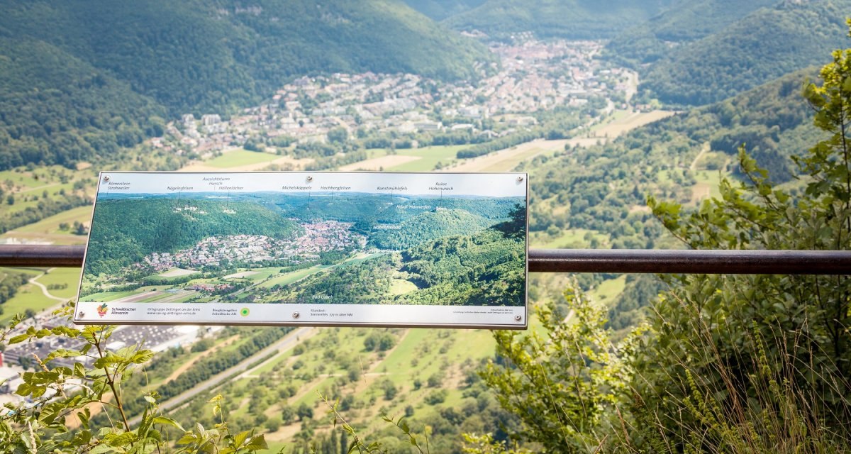 Aussichtspunkt mit Blick auf Bad Urach, umgeben von grünen Hügeln. Eine Informationstafel zeigt Details der Landschaft., © hochgehberge Aussichtspunkt mit Blick auf Bad Urach, umgeben von grünen Hügeln. Eine Informationstafel zeigt Details der Landschaft., © hochgehberge