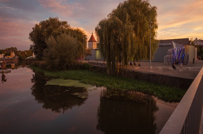 Ein Fluss bei Sonnenuntergang mit B&auml;umen und einem Turm im Hintergrund. Menschen spazieren entlang des Ufers, w&auml;hrend der Himmel in warmen Farben leuchtet., &copy; WTM GmbH Waiblingen