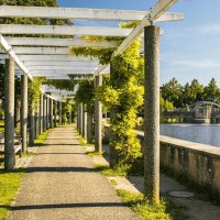 Ein sonniger Weg mit Pergola und Bänken führt entlang eines künstlichen Sees mit Springbrunnen. Grüne Bäume und Pflanzen säumen den Weg., © Stuttgart-Marketing GmbH, Sarah Schmid