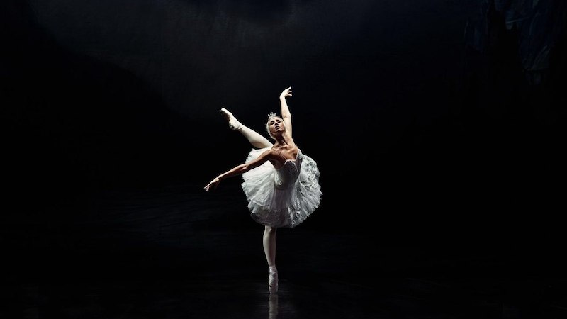 Eine Ballerina in einem weißen Tutu tanzt anmutig auf einer dunklen Bühne, beleuchtet von einem sanften Licht., © Stadthalle Leonberg