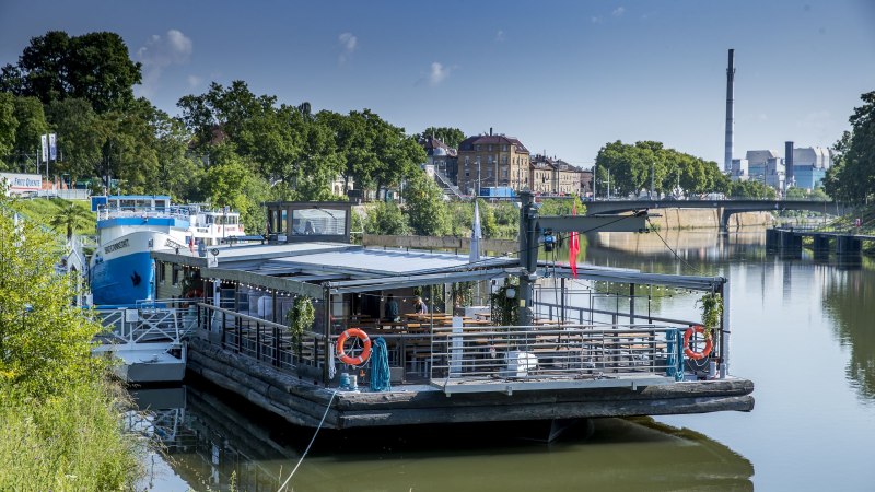 Ein schwimmendes Restaurant auf dem Neckar, umgeben von grüner Vegetation und städtischen Gebäuden im Hintergrund., © all copyrights are reserved by maks richter Ein schwimmendes Restaurant auf dem Neckar, umgeben von grüner Vegetation und städtischen Gebäuden im Hintergrund., © all copyrights are reserved by maks richter