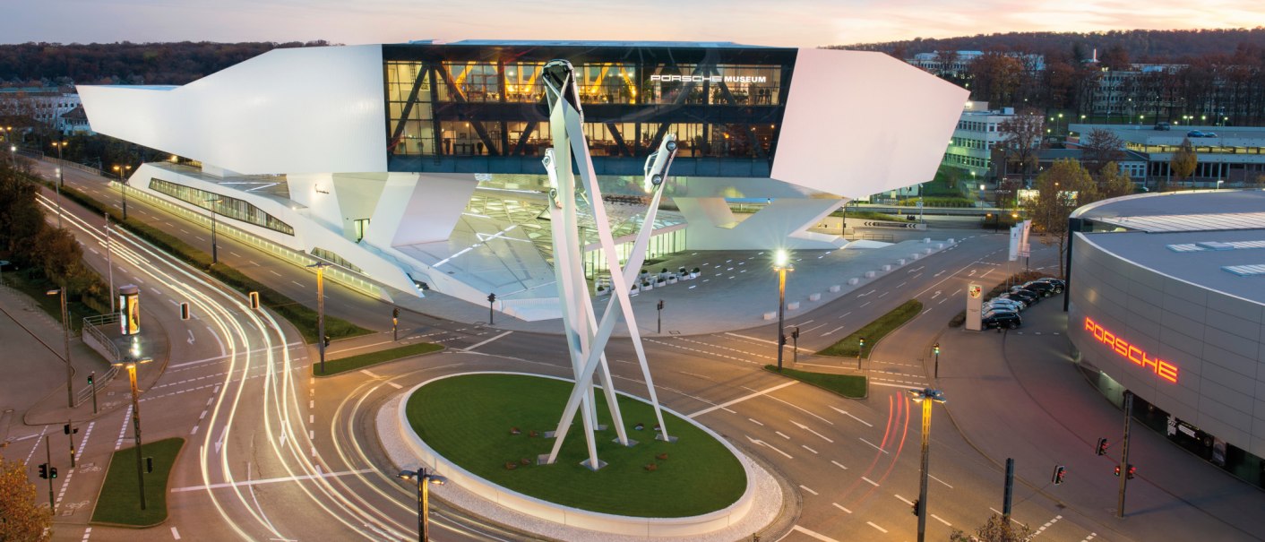 Das Porsche Museum in Stuttgart bei Dämmerung, moderne Architektur mit beleuchteten Straßen und einem Skulpturkreisverkehr im Vordergrund., © Porsche AG Sabine Braun