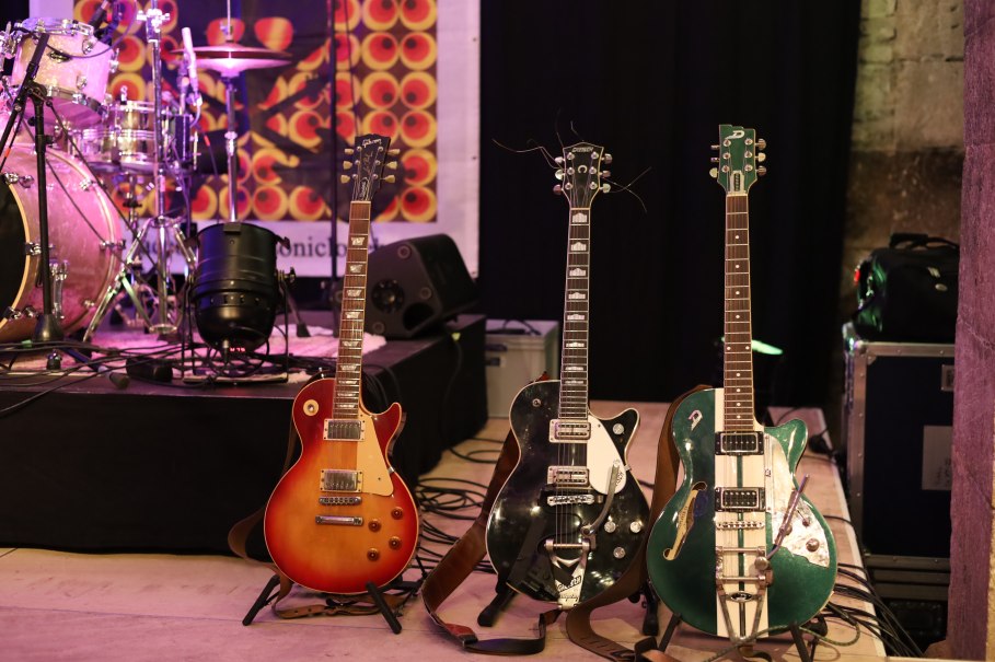 Drei E-Gitarren stehen auf einer Bühne neben einem Schlagzeug. Im Hintergrund ist ein buntes Banner zu sehen., © Stadt Nürtingen Drei E-Gitarren stehen auf einer Bühne neben einem Schlagzeug. Im Hintergrund ist ein buntes Banner zu sehen., © Stadt Nürtingen